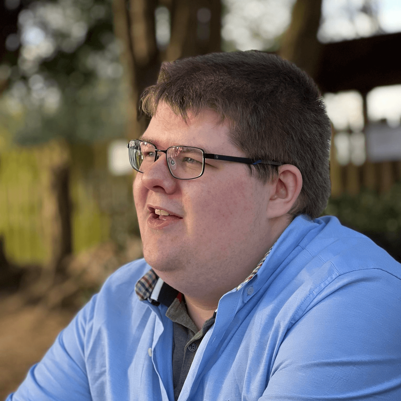 Side-on portrait photo of my face, mouth slightly open smiling wearing glasses with brown hair in a light blue polo shirt